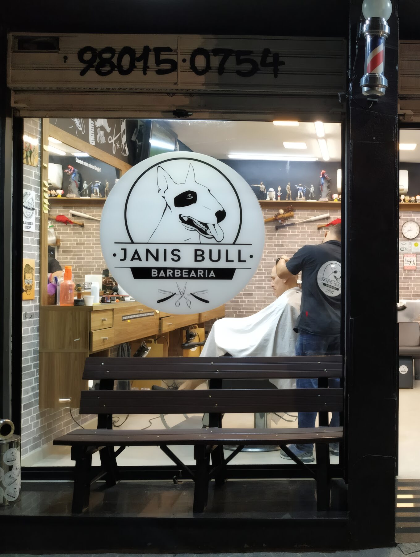 Fachada da Janis Bull à noite: logo do bull terrier iluminado na vitrine, banco de madeira e barber pole aceso